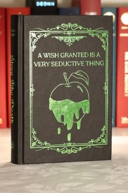 A black hardcover book with a intricate green frame around the words 'A WISH GRANTED IS A VERY SEDUCTIVE THING' and an outline of an apple below it with green substance covering half of it and dripping down.